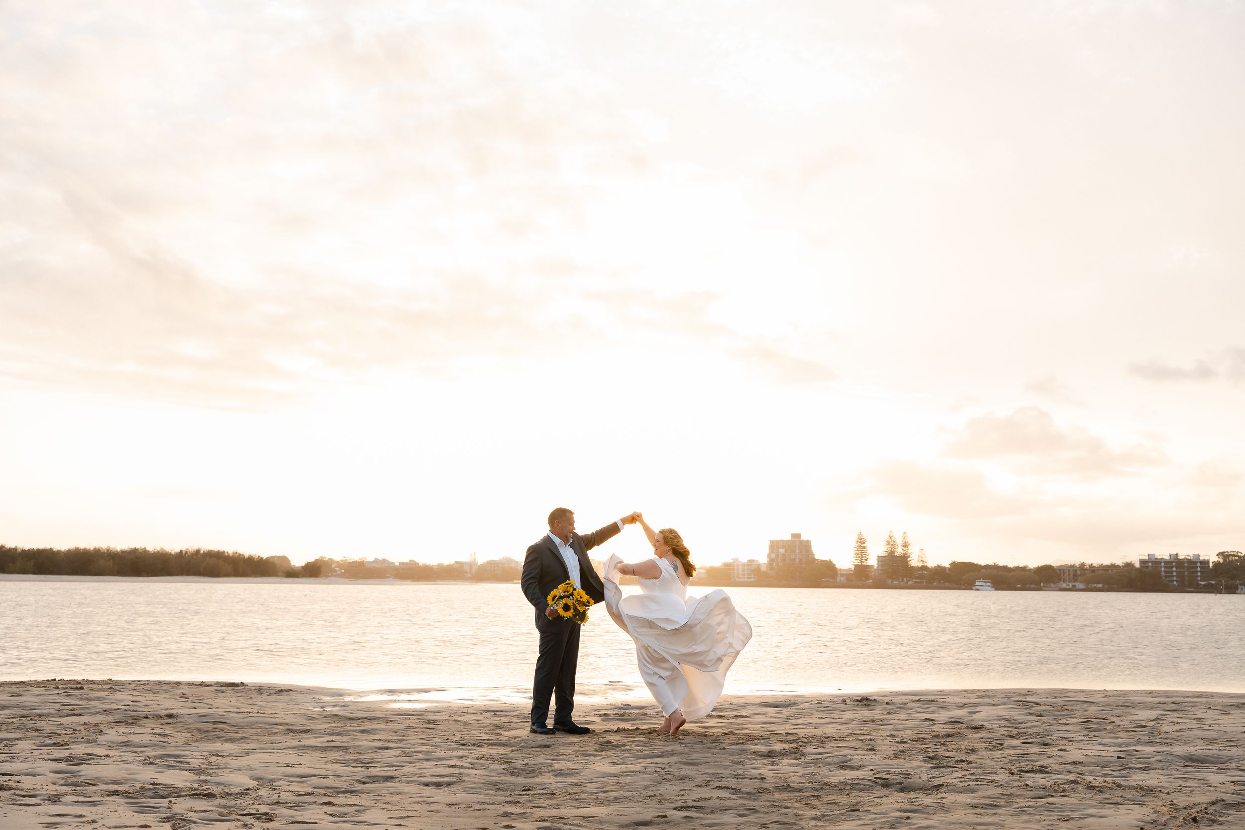 Two silhouettes stand together on a beach at sunset while holding a flowing veil in the wind.
