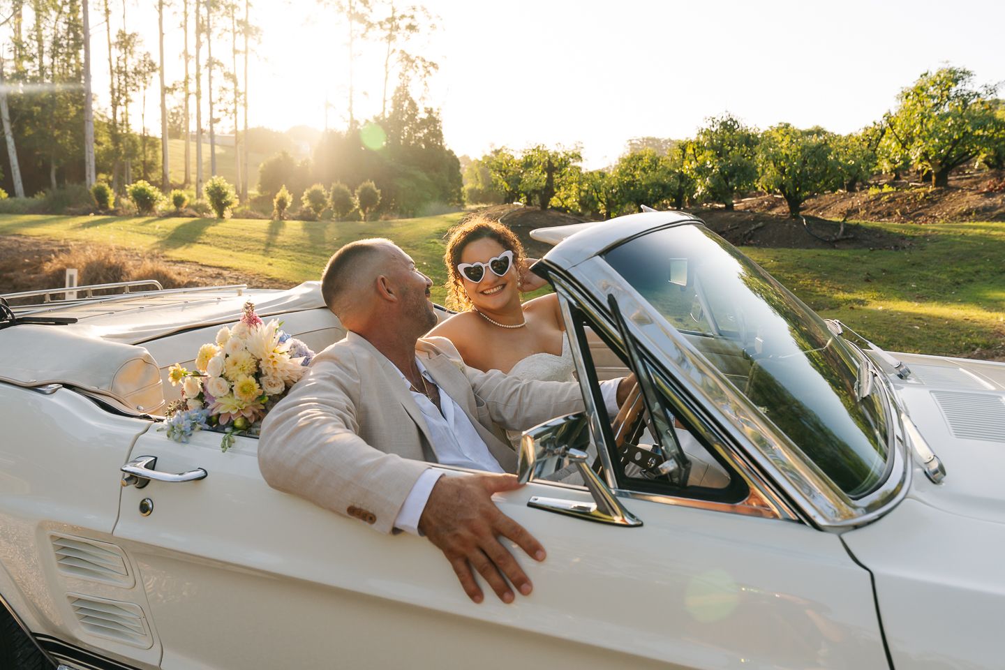 Classic white convertible car cruising along a scenic road at sunset with golden sunlight streaming through trees.
