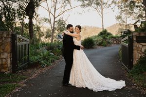 Couple in wedding attire dancing and embracing on a scenic outdoor path surrounded by trees and greenery.