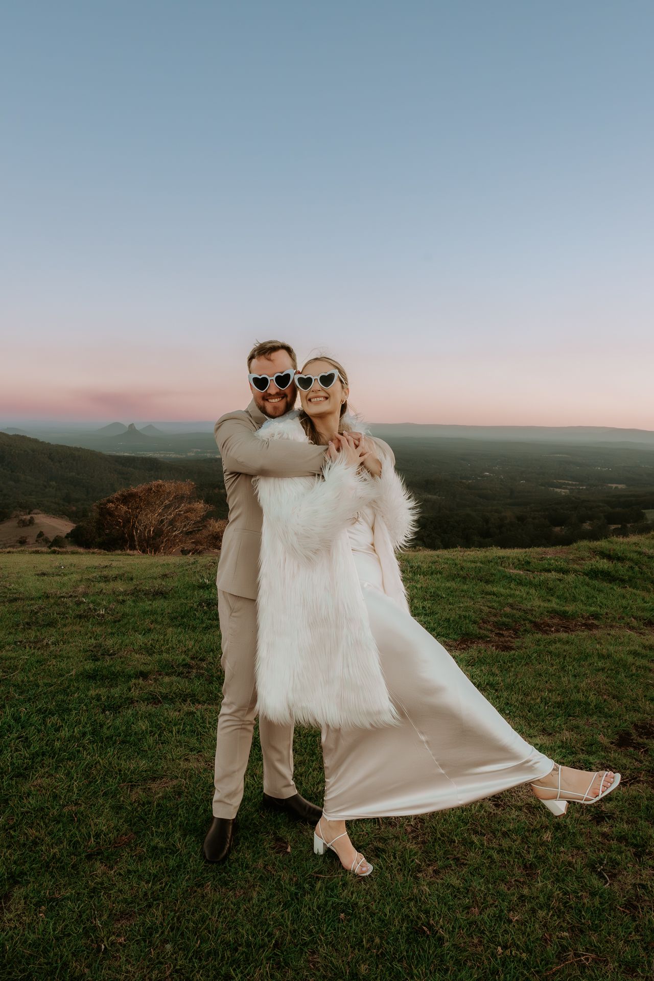 Playful wedding couple in sunglasses posing on hilltop during sunset.