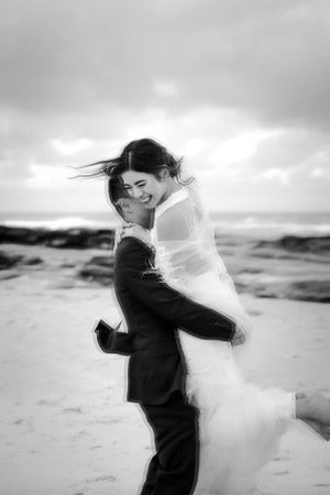 Joyful black and white beach wedding portrait with couple embracing against cloudy sky.