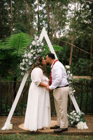 Wedding couple shares intimate moment under white triangle arch decorated with flowers in fern-filled forest.