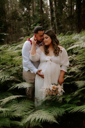 Romantic maternity portrait of couple embracing among ferns in a mystical forest setting.