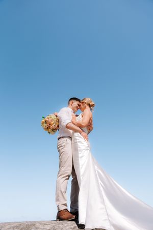 A couple in wedding attire embraces on a rocky outcrop against a bright blue sky.