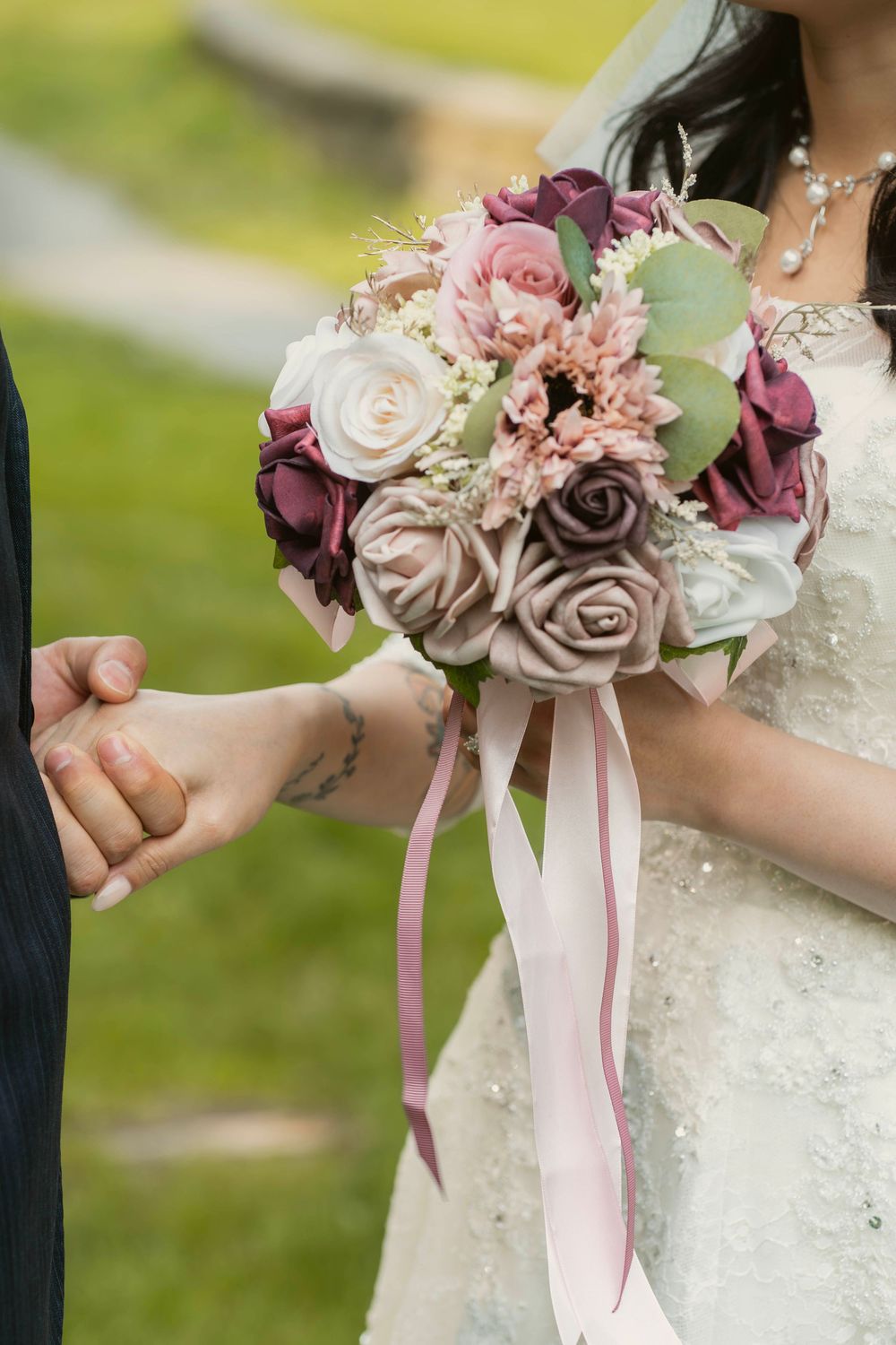 A delicate bridal bouquet featuring dusty rose and mauve flowers with trailing pink ribbons held against a white dress.