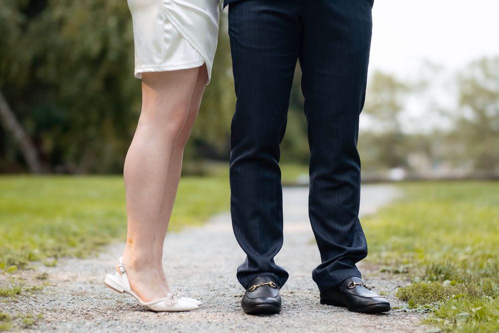 Feet of a bride in white shoes and groom in black dress shoes standing together on a grassy path.