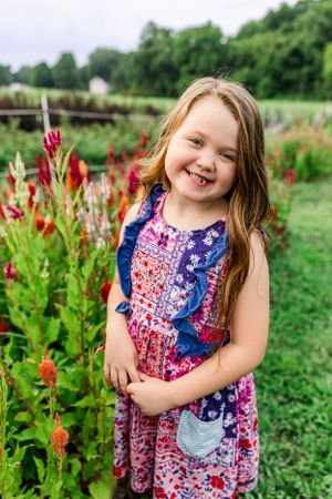 Young girl in floral dress poses by colorful red garden flowers on sunny summer day.