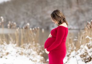 Expectant mother in red dress stands among winter cattails.