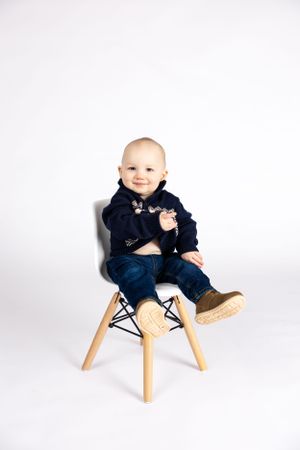 Smiling baby in navy outfit sits on modern wooden chair against white background.