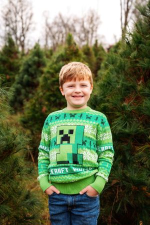 Young boy in green Minecraft Christmas sweater smiles among evergreen trees.