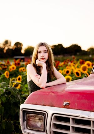 Person leaning on red vintage truck hood surrounded by vibrant sunflowers at sunset.