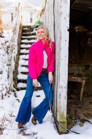 Woman in bright pink jacket poses by snowy stairs near rustic building.