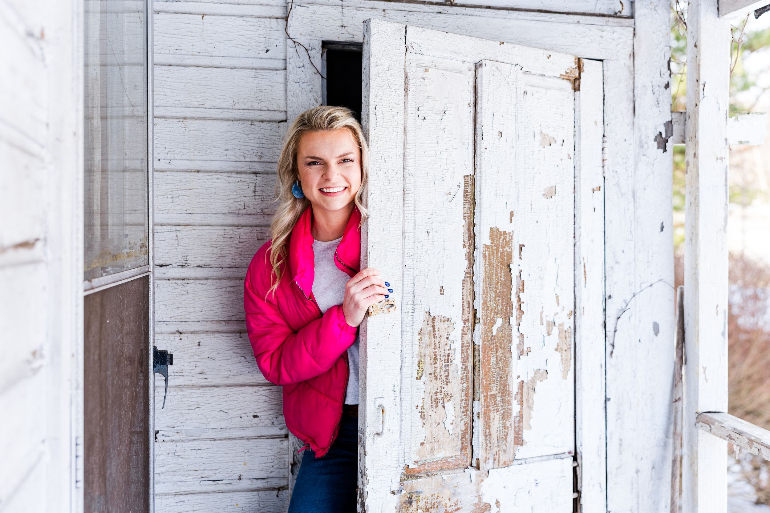 Woman in vibrant pink sweater peeks around weathered white barn door.