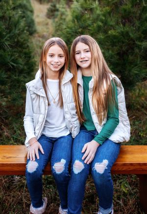 Two teenagers in casual winter wear sit together on wooden bench surrounded by pine trees.