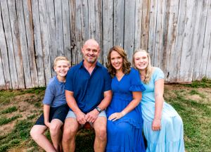 Casual family portrait of four against weathered barn wood background wearing coordinated blue attire.