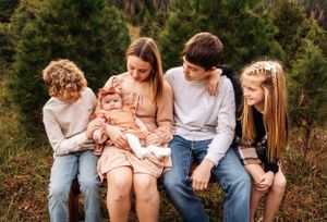 A family of five sits together on a wooden bench surrounded by evergreen trees.