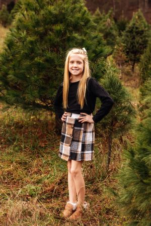 A young person poses in a plaid skirt and black sweater among Christmas trees.