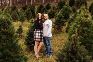 A couple poses together among evergreen trees wearing coordinated casual outfits.