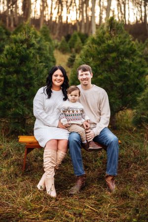A family sits together on a wooden bench surrounded by evergreen trees at sunset.