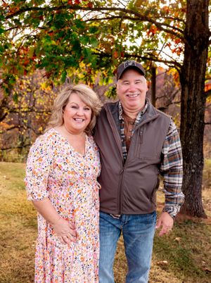 A couple stands together smiling under autumn trees in casual outdoor attire.