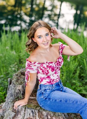 Young woman in floral top poses on tree stump in natural outdoor setting.