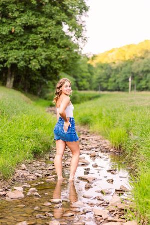 Young woman in denim dress stands playfully in shallow creek on summer day.