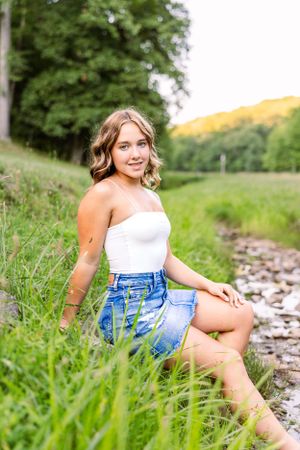 Summer portrait of someone in a white top and denim shorts sitting by a creek surrounded by lush greenery.