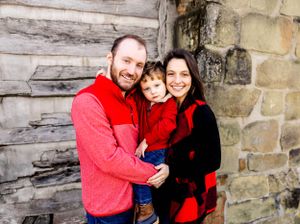 Family of three in coordinating red outfits pose against rustic stone wall.