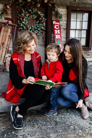 Three people in red attire read book together on rustic porch with Christmas decorations.