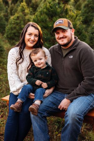 Family sits together on wooden bench surrounded by evergreen trees at Christmas tree farm.