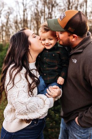 A warm family moment captured outdoors with kisses and hugs among evergreen trees.