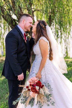 Romantic wedding couple share intimate moment under willow tree with red bouquet.
