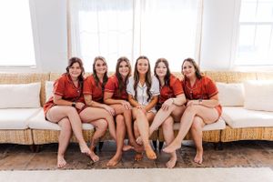 Bridesmaids in matching red robes sit together on beige couch.
