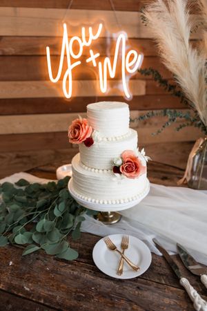 Elegant three-tier white wedding cake with pink roses under neon 'You + Me' sign.