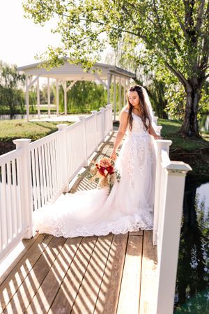 Bride in lace dress poses on white bridge with red bouquet.