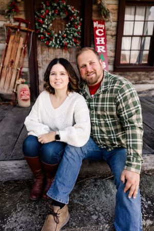 Couple sitting on rustic porch with Christmas decorations and wreath in background.