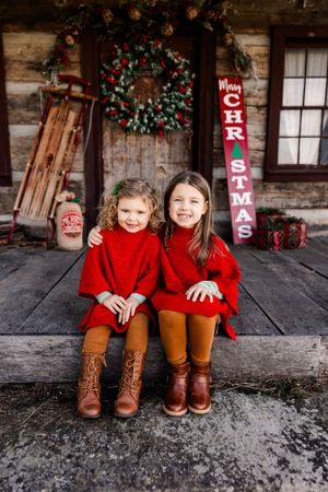 Two children in matching red sweaters sitting on rustic porch with Christmas decorations.