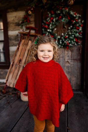 Young child in red sweater smiling against rustic cabin with Christmas wreath.