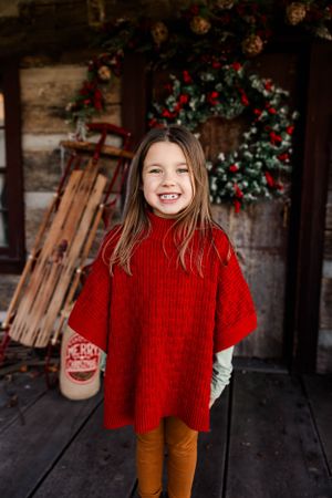 Girl in red sweater smiling in front of cabin with Christmas wreath.