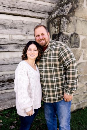 Couple posing against weathered log cabin wall in casual winter attire.