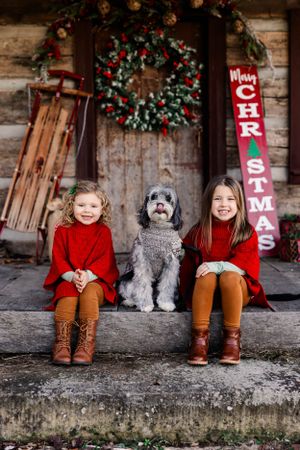 Two children in red sweaters sitting with grey dog on cabin porch with Christmas decor.