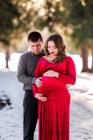Winter maternity photo with red dress against snowy forest backdrop.