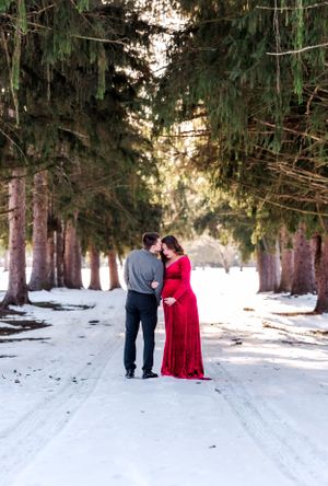 Couple stands together on snowy path between tall evergreen trees.