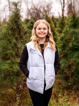 A person in a white vest and black sweater stands smiling among evergreen trees.