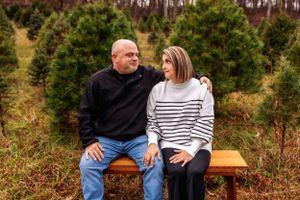 A couple sits close together on a wooden bench surrounded by evergreen trees.
