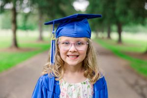 Graduate in blue cap and gown smiling on tree-lined path.