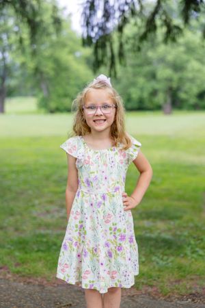 Young girl in floral dress standing on path under trees in summer garden setting.