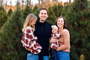 A cheerful group poses together wearing coordinated holiday outfits among evergreen trees at a Christmas tree farm.