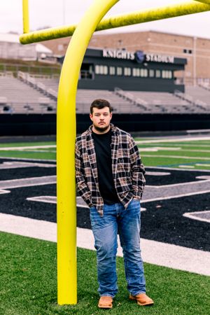 Player stands by yellow goal post on football field with stadium bleachers in background.