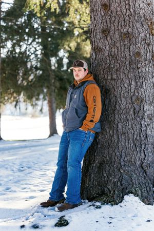 Person in brown Carhartt jacket and jeans leaning against large tree trunk in snow.